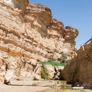 Am Fuße einer geschichteten Felsenschlucht stürzt ein kleiner Wasserfall in ein Wasserbecken, zu dem Steinstufen hinabführen.
