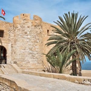 Eine Steinfestung mit einer tunesischen Flagge thront an einem Sandstrand mit Palmen und dem Meer.