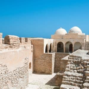 Eine alte Steinfestung mit zwei Kuppeln und gewölbten Eingängen überblickt das Meer unter einem klaren blauen Himmel.