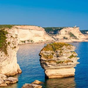 Eine große, geschichtete Felsformation liegt im blauen Meer neben weißen Küstenklippen unter einem klaren Himmel.