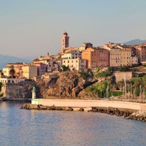Eine farbenfrohe Küstenstadt mit einem markanten Glockenturm liegt auf einer felsigen Klippe über dem Meer, mit einem kleinen Leuchtturm auf einer Mole.
