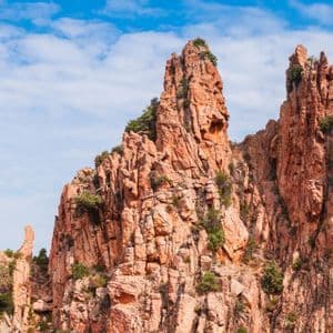 Gezackte, rot-orange Felsformationen mit spärlicher grüner Vegetation stehen vor einem strahlend blauen Himmel mit vereinzelten weißen Wolken.