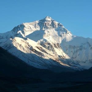 Una grande cima montuosa coperta di neve è illuminata dal sole contro un cielo azzurro limpido, con una valle scura e in ombra in primo piano.