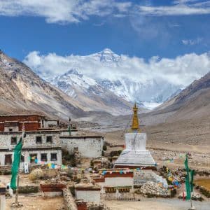 Un villaggio tibetano e uno stupa bianco in una valle ai piedi del Monte Everest innevato sotto un cielo azzurro con nuvole.