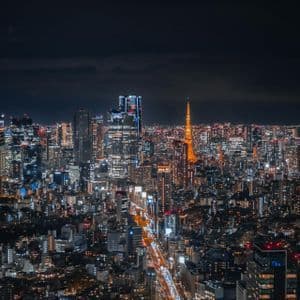 Un vasto skyline cittadino illuminato di notte, con una torre arancione prominente e scie di luce dal traffico su un'autostrada sottostante.
