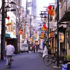 Una persona in bicicletta percorre una stretta strada giapponese fiancheggiata da negozi, insegne e lanterne, con una torre moderna in lontananza.