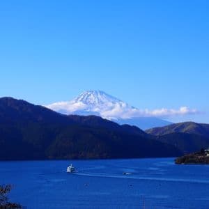 Un traghetto bianco naviga su un lago azzurro, con montagne boscose e il Monte Fuji innevato visibili sullo sfondo.