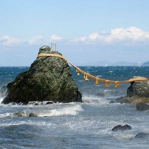 Due grandi faraglioni in un oceano agitato sono collegati da una spessa corda. La roccia più grande è sormontata da un piccolo torii.