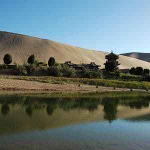 Una pagoda tradizionale asiatica e altri edifici si ergono accanto a un lago calmo alla base di una grande duna di sabbia, riflessi nell'acqua.