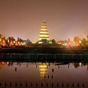 Una pagoda imponente a più livelli è illuminata di notte, la sua luce dorata si specchia nell'acqua calma di una vasca in primo piano.