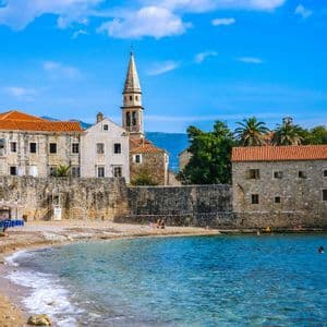 Una storica città costiera con un imponente campanile e edifici in pietra si affaccia su una piccola spiaggia con acque limpide e turchesi sotto un cielo azzurro.