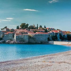Un borgo di pietra con tetti di tegole rosse su una piccola isola rocciosa, ammirato da una spiaggia di ciottoli accanto ad acque calme e turchesi.