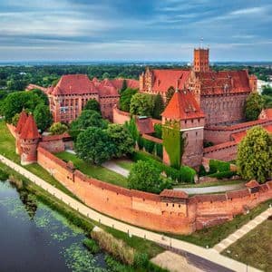 Vue aérienne d'un grand château médiéval en briques rouges avec des tours, entouré d'un mur fortifié et d'une rivière, au milieu d'arbres verdoyants.