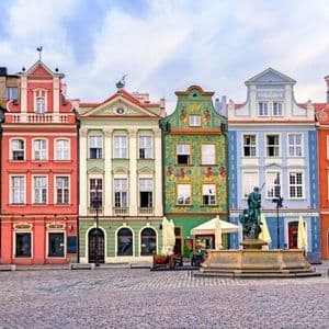 Une rangée de bâtiments colorés et ornés bordent une place de ville pavée avec une fontaine au premier plan.