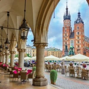 Vue d'une terrasse de café installée sous une colonnade de pierre voûtée, sur une place de ville historique, avec une grande église en briques en arrière-plan.