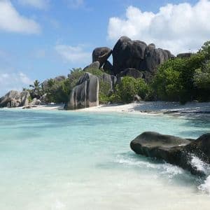 Große graue Felsen säumen einen weißen Sandstrand neben klarem türkisfarbenem Wasser unter einem blauen Himmel mit Wolken.