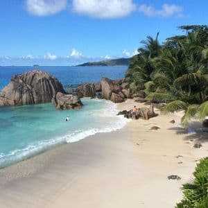 Ein erhöhter Blick auf einen tropischen Strand mit weißem Sand, türkisblauem Wasser, großen Granitfelsen und Palmen unter sonnigem Himmel.
