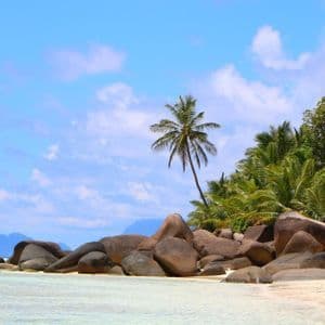 Ein tropischer Sandstrand mit großen, glatten Felsen und üppigen Palmen am klaren Wasser, unter blauem Himmel mit Wolken.