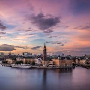 Un paesaggio urbano con un'alta guglia di chiesa su un lungomare, con edifici che si riflettono nell'acqua calma sotto un cielo viola e rosa al tramonto.