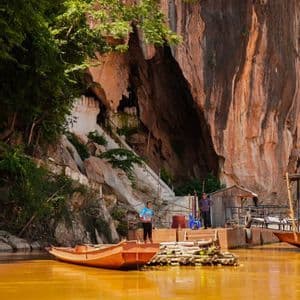 Une pirogue à queue longue est amarrée à un ponton en bois sur une rivière boueuse, devant une grande falaise avec une entrée de grotte.