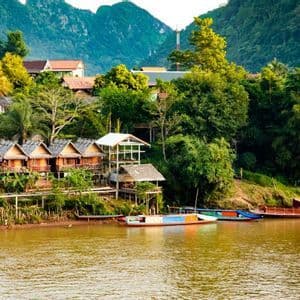 Un village de bungalows en bois sur pilotis est installé au bord d'une rivière, entouré d'une jungle luxuriante et de montagnes, avec plusieurs bateaux amarrés sur l'eau.