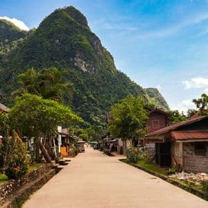 Une rue paisible traverse un village champêtre, bordée de maisons et d'arbres, menant vers une grande montagne boisée sous un ciel bleu.