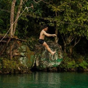 Un homme torse nu en short noir saute dans une rivière verte depuis un rocher dans une jungle luxuriante.