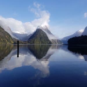 Forested mountains with snow-capped peaks are reflected in the still, clear water of a fjord under a blue sky.