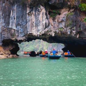 Un viaggio di gruppo WeRoad in piccole barche a remi attraverso un grande arco di roccia naturale su acqua verdastra.