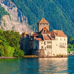A historic stone castle with multiple towers sits on the shore of a lake, at the base of a steep, forest-covered mountain.