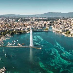 Vista aérea de una gran fuente de agua que brota de un lago turquesa, con una densa ciudad y un puerto deportivo a lo largo de la orilla.