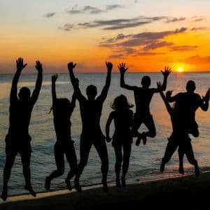 Un voyage de groupe WeRoad en silhouette devant un coucher de soleil orange, sautant ensemble sur une plage au bord de la mer.