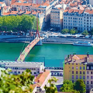 Vue plongeante sur un paysage urbain, avec un pont piétonnier rouge au-dessus d'une rivière turquoise, bordée de bâtiments et d'arbres.
