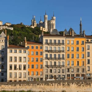 Des bâtiments colorés bordent une rivière, avec une cathédrale blanche sur une colline verdoyante en arrière-plan, sous un ciel bleu clair.