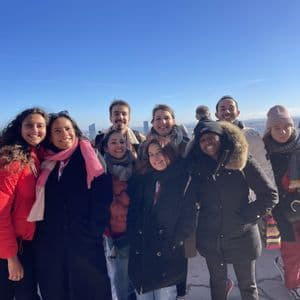 Un groupe WeRoad de neuf personnes souriant pour une photo, vêtues de manteaux d'hiver depuis un point de vue de la ville sous un ciel bleu clair.