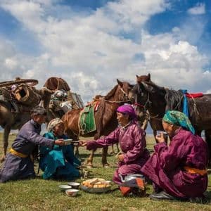 Eine Gruppe Menschen in traditioneller Kleidung teilt eine Mahlzeit auf einer grasbewachsenen Ebene, dahinter stehen ein Lastkamel und mehrere Pferde.
