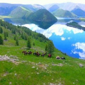 Eine WeRoad Gruppenreise zu Pferd ruht auf einem grünen Hügel mit Blick auf einen ruhigen See, der den Himmel und die Berge widerspiegelt.