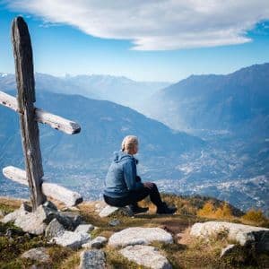 Una persona con trenzas rubias se sienta en un afloramiento rocoso junto a una cruz de madera, con vistas a un amplio valle de montaña.
