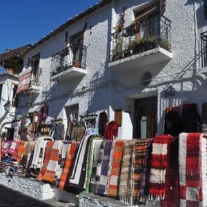 Alfombras tejidas coloridas y ropa cuelgan en exhibición en una calle frente a un edificio blanco bajo un cielo azul claro.