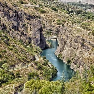 Vista aérea de un río turquesa serpenteando por un cañón rocoso y escarpado, con laderas áridas cubiertas de vegetación y una presa distante.