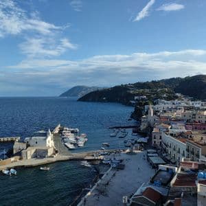 Vista aerea di una colorata cittadina costiera e del suo porto pieno di barche, adagiata su un litorale collinare sotto un cielo azzurro.