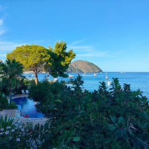 Una piscina circondata da alberi rigogliosi si affaccia su un mare tranquillo costellato di barche e un'isola collinare in lontananza.