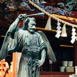 Una statua in bronzo di un Tengu alato, divinità giapponese con un becco prominente, si erge all'esterno di un tradizionale tempio rosso.