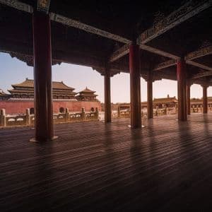 Una vista da una veranda in legno scuro con pilastri rossi, che si affaccia su tradizionali palazzi cinesi con tetti gialli contro un cielo pallido.