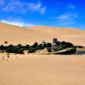 Un'oasi con un lago a mezzaluna e una pagoda tradizionale è incastonata tra vaste dune di sabbia del deserto sotto un cielo azzurro brillante.