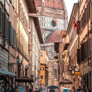 Una vista su una stretta via assolata di una città storica, con persone che passeggiano e una grande cupola di cattedrale dalle tegole rosse sullo sfondo.