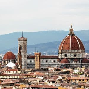 Una cattedrale con un'ampia cupola di tegole rosse e un campanile si erge sopra un paesaggio urbano di tetti in terracotta, con le montagne sullo sfondo.