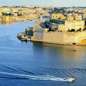 A white motorboat speeds across blue water, creating a wake, with a large stone fortress and coastal city in the background.