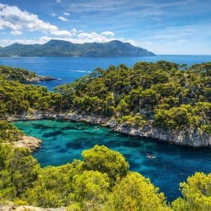 Una vista panoramica di una cala turchese circondata da pini verdi e coste rocciose, con montagne al di là del mare.