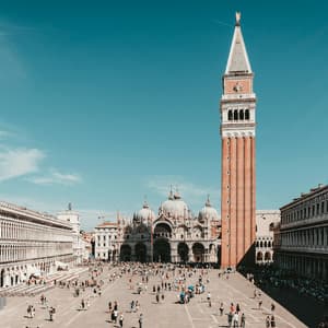 Un día soleado sobre una animada plaza histórica europea llena de gente, con un alto campanario de ladrillo y una gran basílica abovedada.
